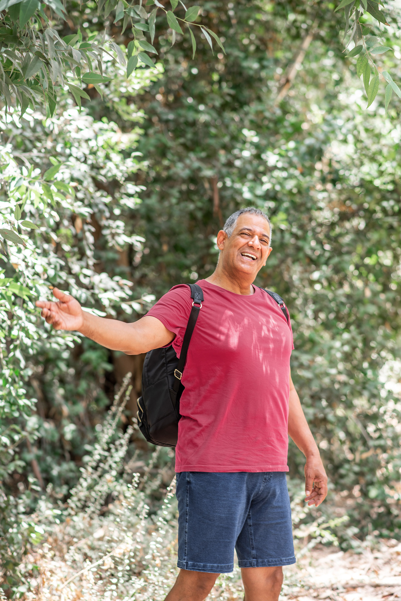Portrait of a happy activity athlete senior man carrying a backpack, enjoying beautiful view. Healthy old man on a hiking trip. Male tour guide in the forest doing a hand inviting gesture.